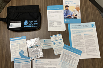 The image shows an "Aware in Care" kit from the Parkinson's Foundation, containing informational pamphlets, forms, and a carrying case designed to help people with Parkinson's disease stay safe in the hospital.