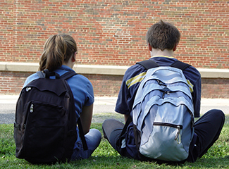 Two students with their backpacks sitting next to each other on the grass.