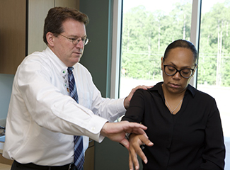 The image shows Dr. Steven Greer examining a patient's arm in a medical office.