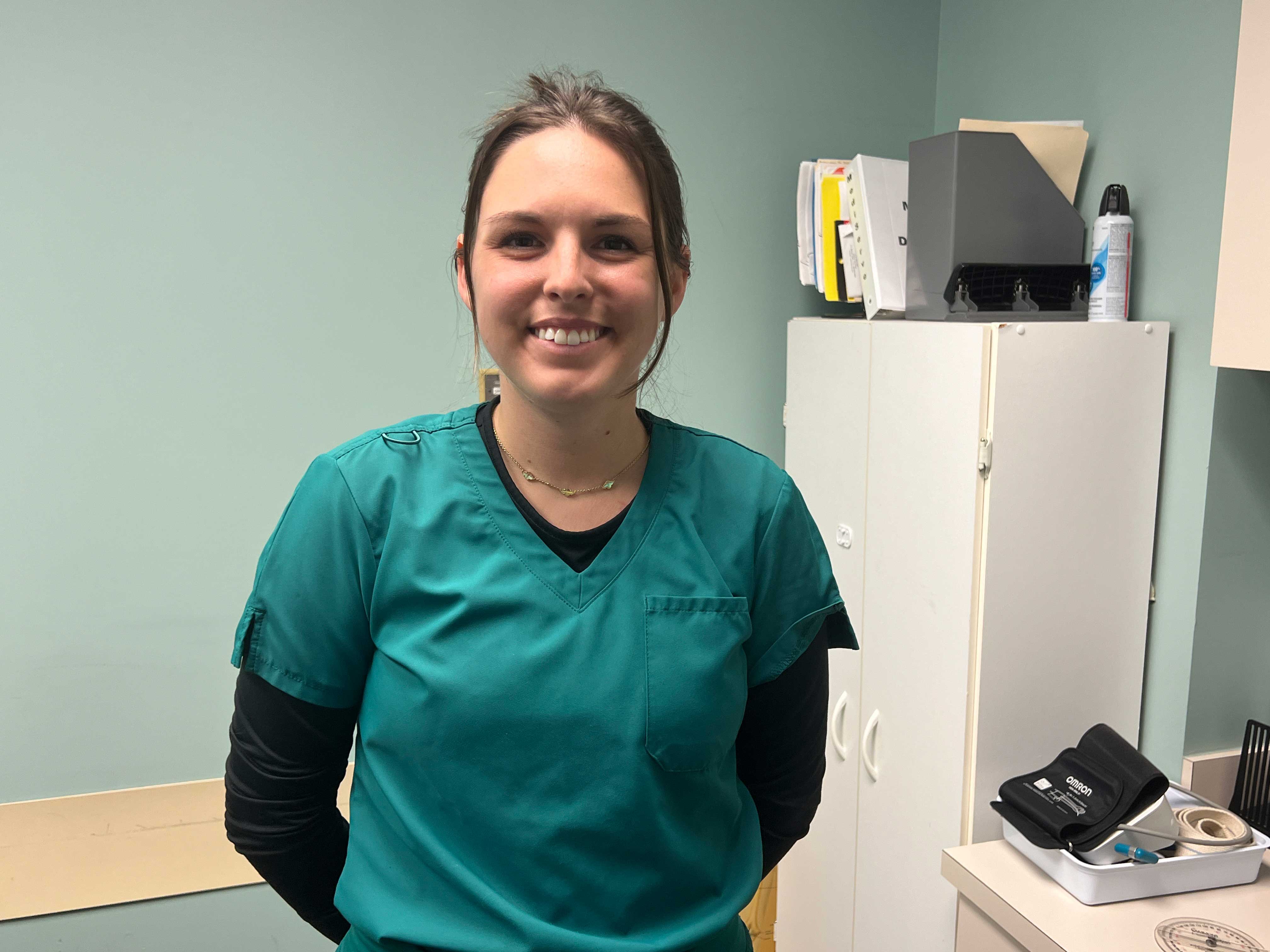 A smiling woman in teal medical scrubs, standing in a clinical office.