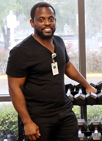 A smiling man in black T-shirt standing next to a rack of dumbbells in a gym or fitness facility.