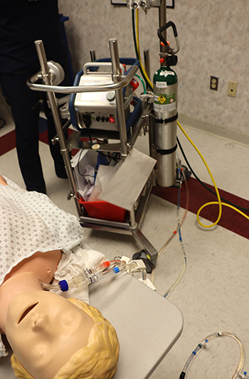 A medical training setup with a mannequin patient connected to an ECMO (extracorporeal membrane oxygenation) machine and oxygen tank.