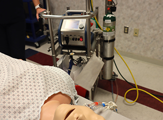 A medical training setup with a mannequin patient connected to an ECMO (extracorporeal membrane oxygenation) machine and oxygen tank.