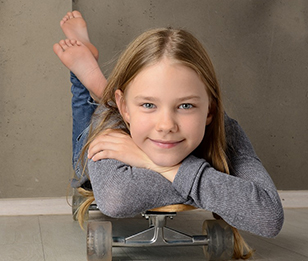 A young girl is lying on a skateboard, smiling at the camera.