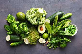 A variety of fresh green vegetables and fruits, including broccoli, avocado, cucumbers, basil, peas, and leafy greens.