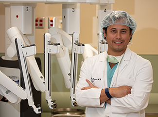 A smiling Dr. Andres Montes, an OB/GYN in surgical attire standing in front of a robotic surgical system.