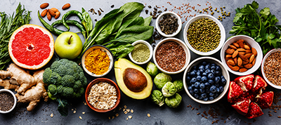 The image shows a variety of fresh fruits, vegetables, nuts, and seeds arranged on a surface, representing healthy nutrition for heart health.