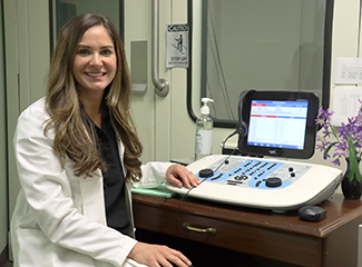 Audiologist Rachel Goldsmith in a white lab coat is sitting at a desk with audiology equipment.