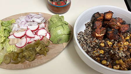 A quinoa bowl with roasted sweet potatoes, corn, and beans, alongside a wooden board with sliced radishes, onions, jalapeños, avocado, and lettuce.