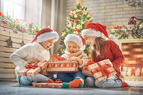 Three children wearing Santa hats are happily opening Christmas presents in a festive, decorated room with a Christmas tree.