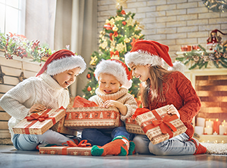 Three children wearing Santa hats are happily opening Christmas presents in a festive, decorated room with a Christmas tree.