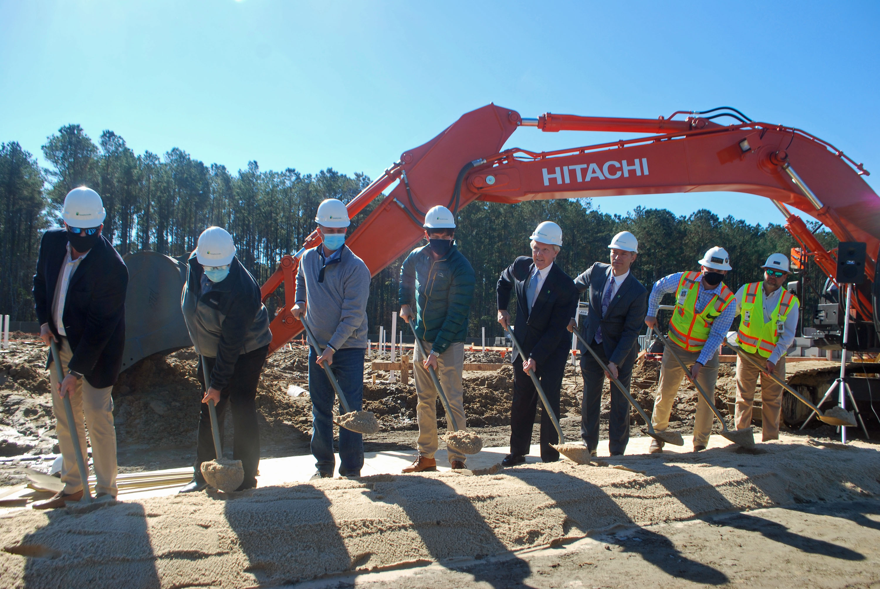 A group of people from RICHMOND HILL - St. Joseph’s/Candler participate in a groundbreaking ceremony, shoveling dirt at the new Urgent Care facility construction site with an Hitachi excavator in the background.
