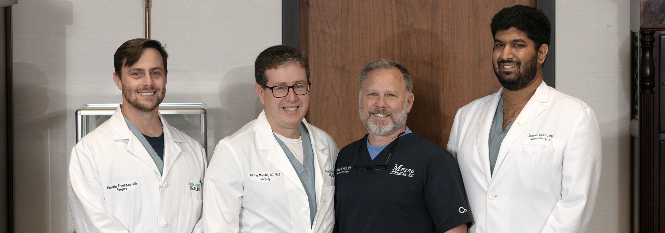 Four male surgeons, three wearing white lab coats and one in navy blue scrubs, standing together and smiling.